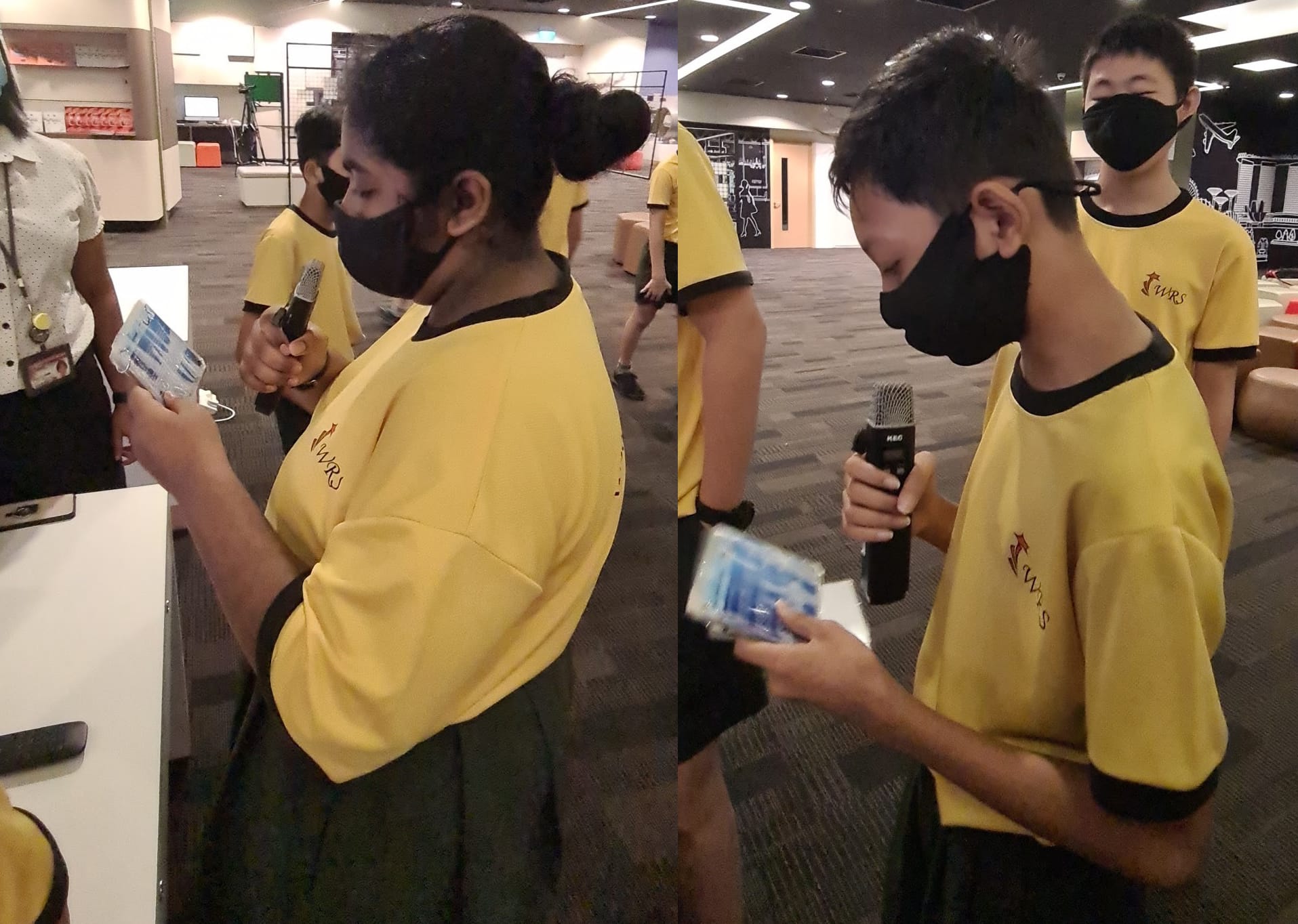 Two students in yellow uniforms and black masks hold microphones and cards indoors with others in the background.